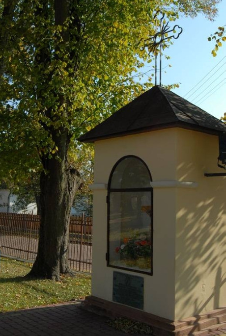 Współczesne kolorowe zdjęcie. Ukazuje kapliczkę przydrożną. Za nią drzewo i ogrodzenie. Jest to kapliczka z tablicą pamiątkową poświęconą m.in. odsieczy wiedeńskiej w Wielkich Oczach. 
