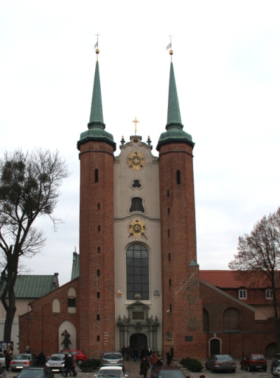 Zdjęcie współczesne, kolorowe. Przedstawia fasadę kościoła. Charakterystyczne dwie wysokie, strzeliste, ceglane wieże. Część środkowa jasna z portalem wejściowym i dużym oknem. Przed świątynią ludzie i zaparkowane samochody. Jest to Archikatedra Oliwska pw. Trójcy Świętej.