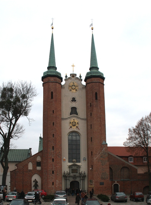 Zdjęcie współczesne, kolorowe. Przedstawia fasadę kościoła. Charakterystyczne dwie wysokie, strzeliste, ceglane wieże. Część środkowa jasna z portalem wejściowym i dużym oknem. Przed świątynią ludzie i zaparkowane samochody. Jest to Archikatedra Oliwska pw. Trójcy Świętej.