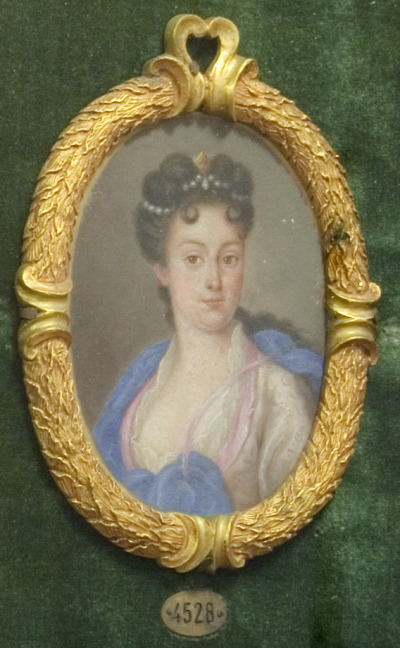 Miniatura portretowa Teresy Kunegundy Sobieskiej. Młoda kobieta w jasnej sukni z dużym dekoltem, na który opada błękity szal. Kobieta ma misternie upiętą fryzurę, zdobioną perłami. Miniatura oprawiona w dekoracyjną, złotą ramę. 
