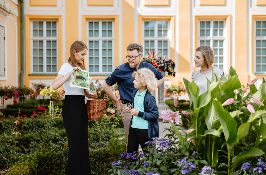 Scena przed pałacem. Na alejce, wśród rabat kwiatowych stoi trójka młodych ludzi i chłopiec. Edukatorka trzyma w jednej ręce kosz, w drugiej kartkę. Pokazuje coś chłopcu.