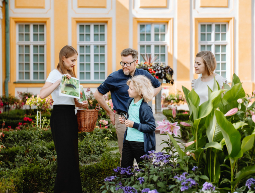 Scena przed pałacem. Na alejce, wśród rabat kwiatowych stoi trójka młodych ludzi i chłopiec. Edukatorka trzyma w jednej ręce kosz, w drugiej kartkę. Pokazuje coś chłopcu.
