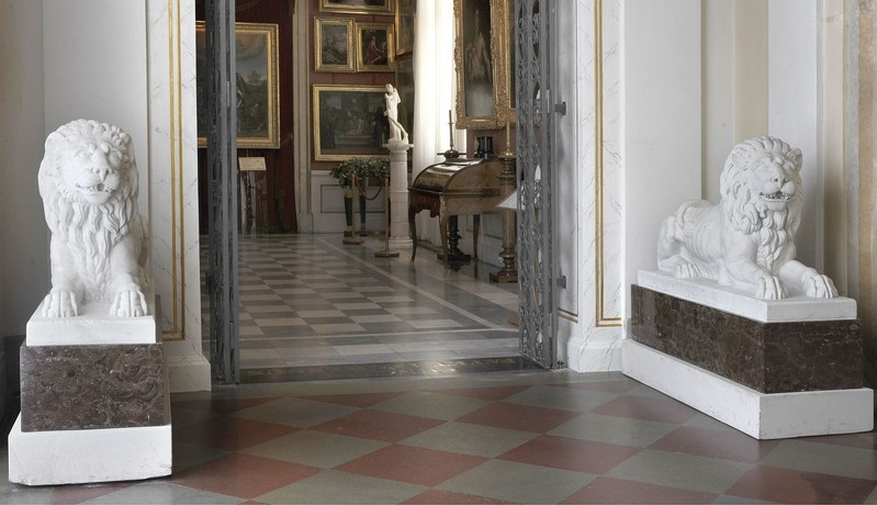 Widok na wnętrze pałacowe. Na pierwszym planie, po bokach drzwi wejściowych do drugiej sali - dwie rzeźby leżących lwów. W głębi sekretarzyk i galeria z obrazami.